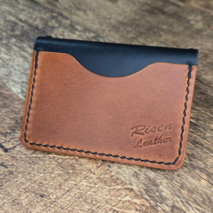 Brown leather wallet with black top section on a wooden surface, featuring 'Risen Leather' branding.