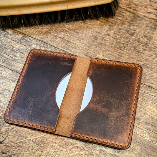 Brown leather cardholder with strap on a wooden surface, next to a brush.
