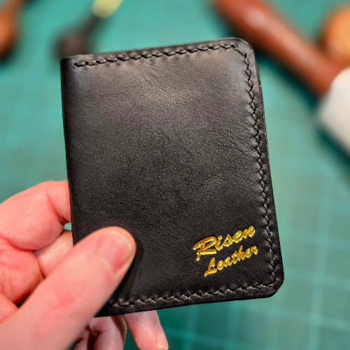 Black leather wallet with 'Risen Leather' branding held by a hand on a green cutting mat.